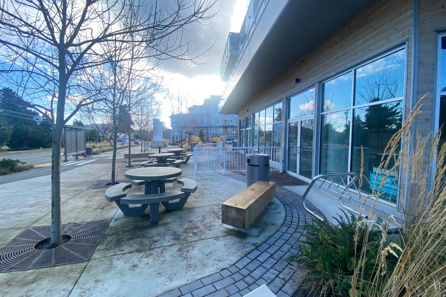 The exterior of Cordova Bay Shopping Center, a commercial property, designed by Alan Lowe Architecture with commercial spaces on the first level, and seating and planted trees out front of them.