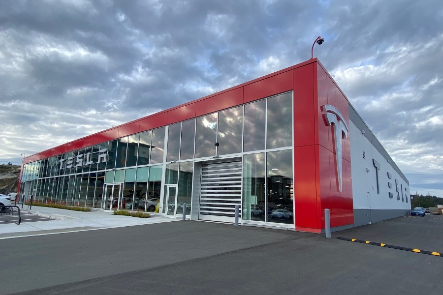 The exterior of Tesla Dealership, a commercial property, designed by Alan Lowe Architecture with large glass front, red and grey siding and large signage.